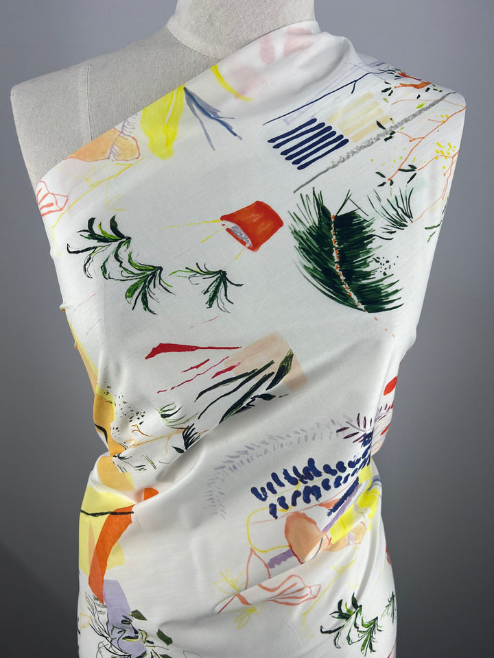 A close-up of a mannequin draped in Super Cheap Fabrics' Designer Cotton - Holiday - 136cm with an abstract, colorful pattern. The design includes various shapes and elements in green, yellow, red, and blue. The multi-use fabric wraps elegantly against the plain gray background.