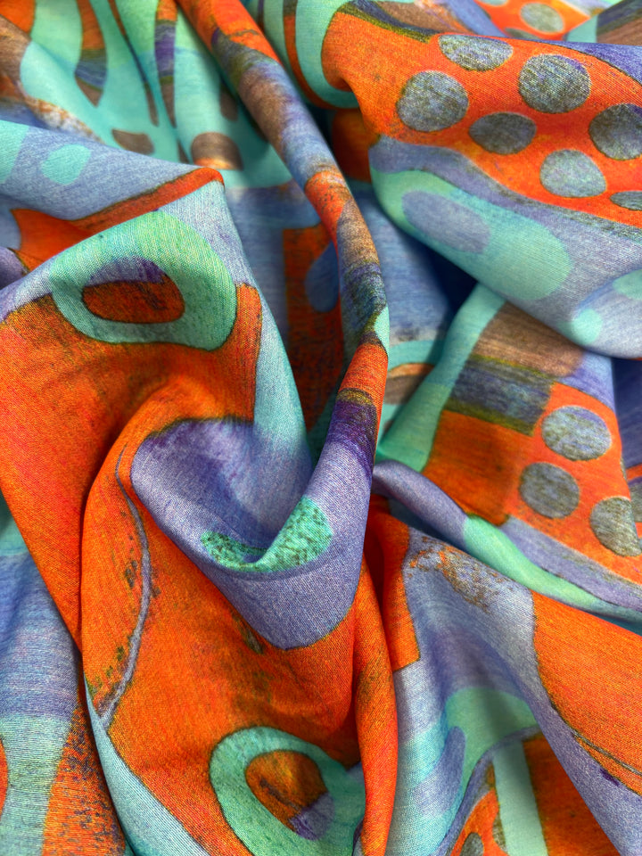 A close-up view of Super Cheap Fabrics' Designer Cotton - Fossil - 150cm featuring a vivid abstract pattern. The design includes bright orange, teal, and purple colors with shapes like circles and curved lines. Perfect for children's clothing, the fabric appears soft and slightly crumpled, showcasing intricate details and vibrant hues.