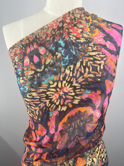 A vibrant, multicolored fabric with an abstract, floral-like pattern is draped over a mannequin. Perfect for fancy dresses, this Designer Chiffon - Tribadelic - 150cm by Super Cheap Fabrics features bold splashes of pink, orange, blue, and purple hues, creating a dynamic and eye-catching design while showcasing its flowing quality.