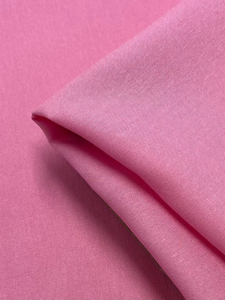 A close-up of the Super Cheap Fabrics' Plain Rayon in Prism Pink, folded at the top right corner to reveal the smooth texture and slight sheen of the material. The fabric, measuring 145cm wide, has a rich, uniform pink color and appears soft, lightweight, and flexible.