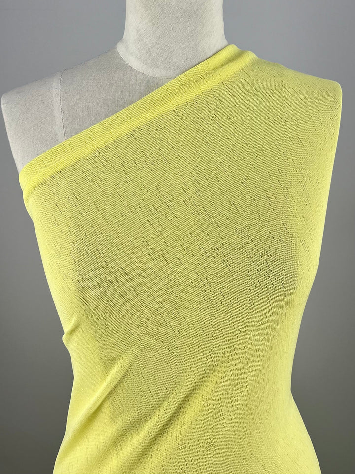 A dress form displays a one-shoulder garment made of the Lightweight Slub Jersey in Lemon from Super Cheap Fabrics. The comfortable stretch material features a subtle, textured knit pattern, and the garment drapes elegantly across the mannequin, leaving one shoulder and arm exposed. The background is a plain, neutral gray.