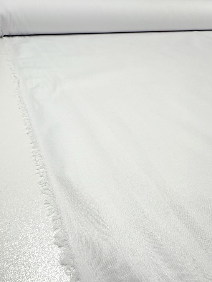 A roll of Stretch Cotton - White from Super Cheap Fabrics, measuring 125 CM in width, unfurled on a smooth surface. The material has a slight fringe along one edge, indicating it has been cut from a larger piece. This lightweight stretch cotton fabric showcases a fine, even texture.