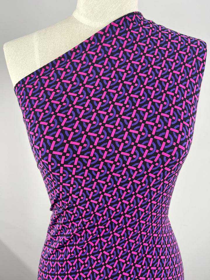 A mannequin wears an asymmetrical dress with a one-shoulder design in medium-weight fabric. The dress features a vibrant pattern of interlocking pink and purple geometric shapes on a dark purple background, made from Printed Lycra - Loopholes - 150cm by Super Cheap Fabrics. The material appears slightly gathered at the waist.