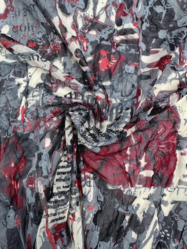 A detailed view of the lightweight Printed Mesh - Read - 120cm from Super Cheap Fabrics, showcasing a collage-like pattern. The design displays irregular red, gray, and black abstract shapes accompanied by text fragments. This soft fabric has a slight tie-dye appearance and an appealing crumpled texture.