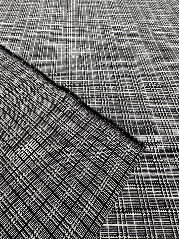 A close-up view of the textured, medium-weight Textured Bengaline - Plaidly fabric from Super Cheap Fabrics, showcasing its checkered pattern in black, gray, and white. A small fold or overlap reveals a subtle variation in the intricate and geometric design beneath this versatile textile. The fabric measures 150cm in width.