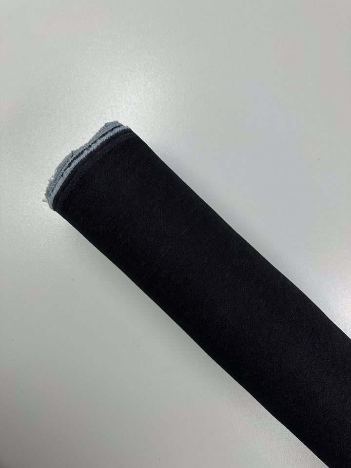 A roll of Super Cheap Fabrics' Lightweight Denim - Black - 135cm is lying on a smooth, light-colored surface. The fabric appears to be tightly wound with a uniform texture and slightly frayed edges.