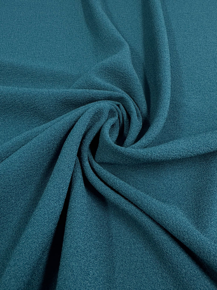 A close-up image of Designer Crepe - Teal - 145cm fabric from Super Cheap Fabrics, gathered in loose folds to create a circular swirl pattern in the center. The fine texture and soft appearance of this medium weight polyester crepe fabric are clearly visible, showcasing its smooth and somewhat matte finish. Perfect for elegant dress material.