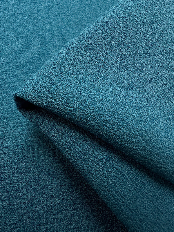 Close-up of two slightly folded pieces of Designer Crepe - Teal from Super Cheap Fabrics, showcasing a finely woven yet somewhat rough surface. The fabric layers overlap, emphasizing the material's thickness and rich teal color, making it ideal for fancy dresses.