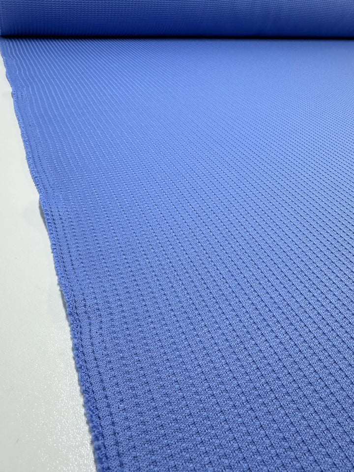 A roll of Super Cheap Fabrics' Textured Knit - Ultramarine - 155cm is laid out, showcasing a close-up of its intricate pattern and ribbed surface. The fabric stretches from the foreground to the background, providing a sense of depth and high material quality.