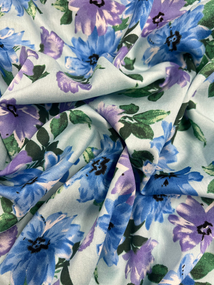 A close-up of Super Cheap Fabrics' Printed Rayon - Watercolour - 145cm, showcasing a light, silky fabric adorned with a beautiful floral pattern. The design highlights various shades of blue and purple flowers with green leaves on a light background. Slightly wrinkled, this versatile fabric creates soft folds and shadows.