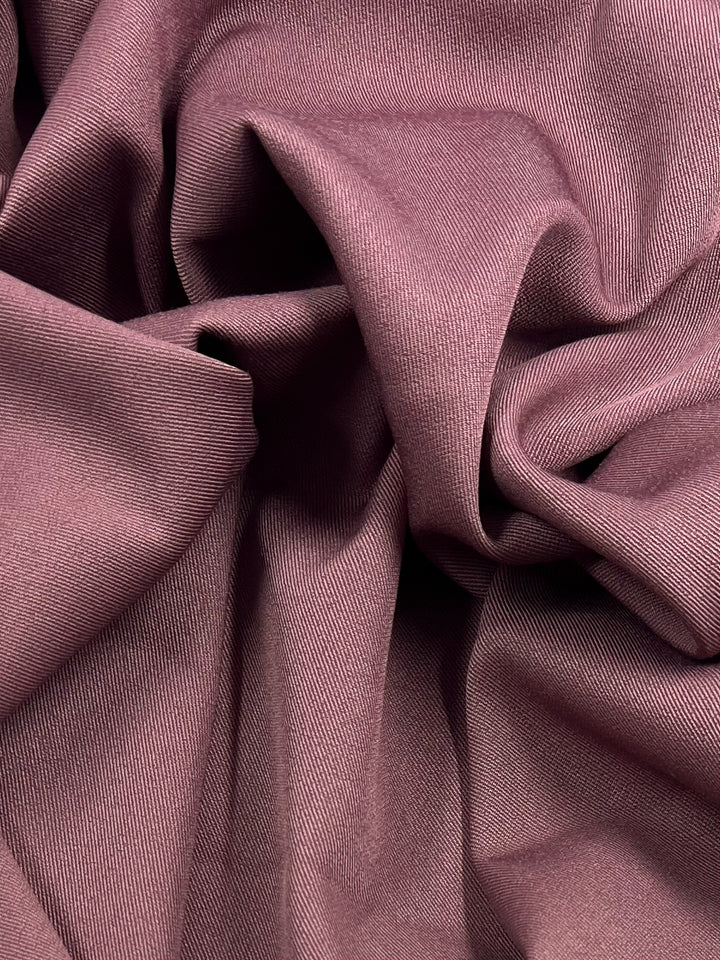 A close-up image of Super Cheap Fabrics' 'Twill Suiting - Mellow Mauve - 150cm' reveals its crumpled, diagonal weave pattern, creating a series of folds and creases that showcase its soft and smooth texture. The hue is a muted pinkish-purple, adding a gentle, elegant appearance to this 100% polyester material.