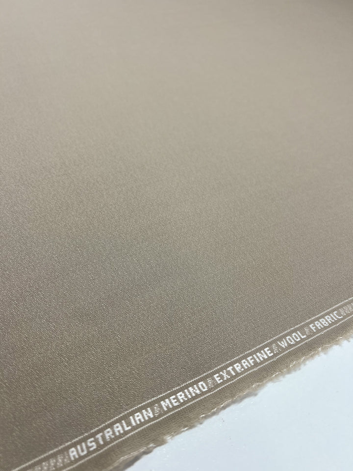 A close-up of the Super Cheap Fabrics' "Merino Wool Suiting - Oxford Tan - 155cm." The edge of the light brown fabric features white text that reads: "AUSTRALIAN MERINO EXTRAFINE WOOL FABRIC." The luxurious texture appears smooth, highlighting the exceptional quality and fineness of the material with excellent breathability.