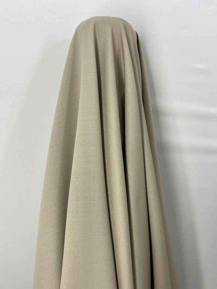 A luxurious fabric from Super Cheap Fabrics, specifically the Merino Wool Suiting in Oxford Tan measuring 155cm, draped over an object against a plain white background. The material hangs down smoothly, creating soft folds and shadows that highlight its excellent breathability and texture.