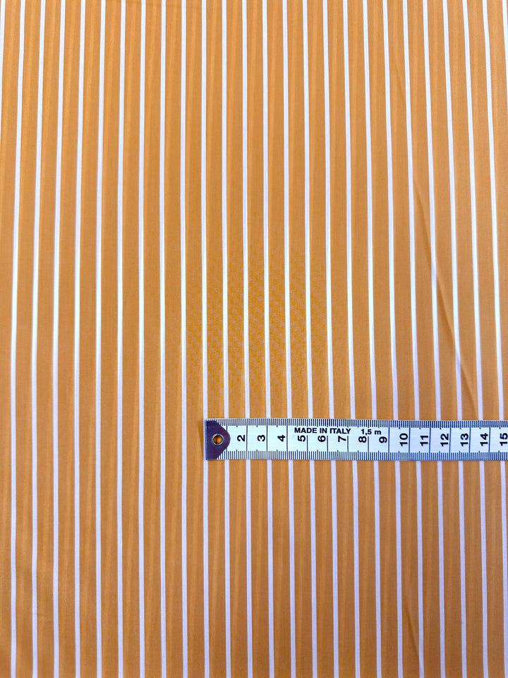 The image features a piece of Polished Cotton Voile - Sunrise - 155cm from Super Cheap Fabrics, made from sustainable deadstock materials with vertical orange and white stripes. A measuring tape in centimeters is placed horizontally across the fabric at the bottom of the image, indicating the scale.