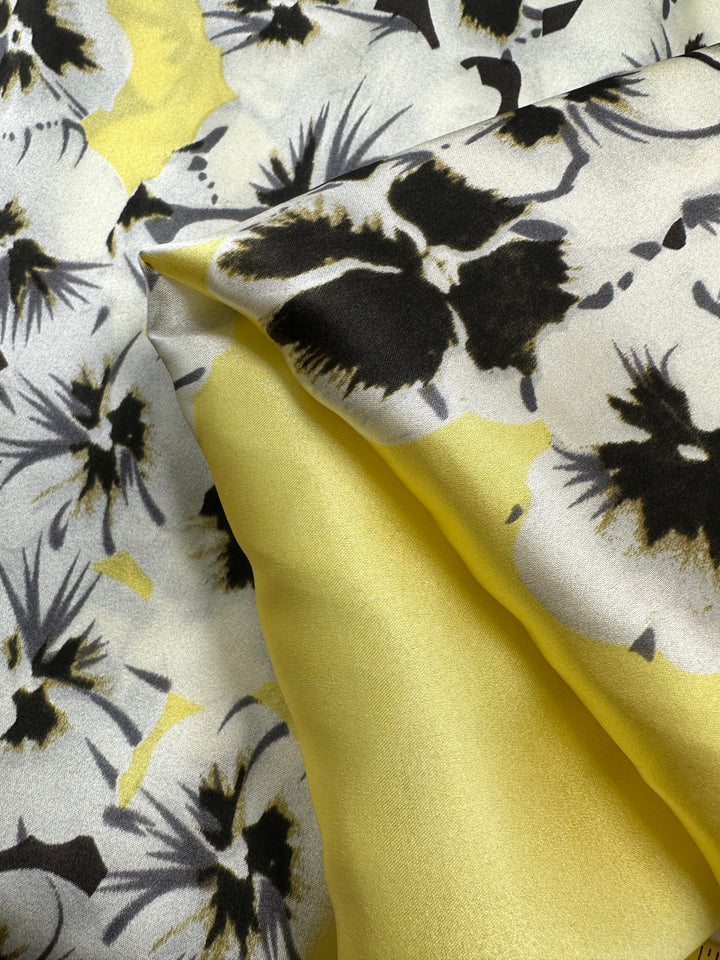 A close-up of the versatile Printed Stretch Silk - Ramona - 100cm by Super Cheap Fabrics showcases a pattern of black and white floral designs on a light background. The fabric also features a smooth, solid yellow section blending into the floral pattern, creating a visually appealing contrast, perfect for high-end fashion attire.