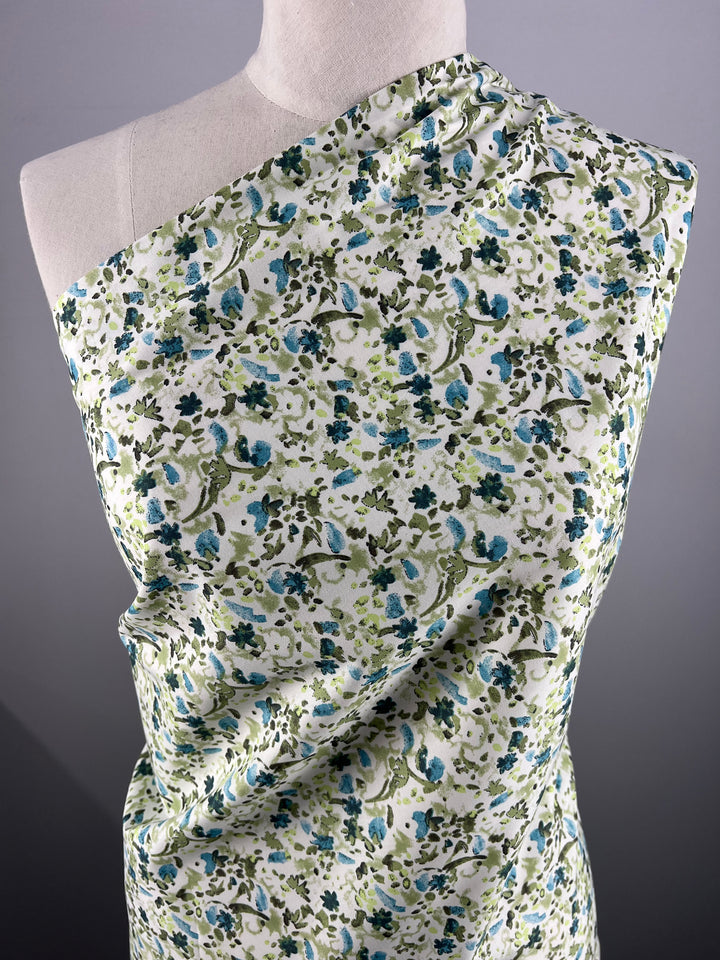 A mannequin draped in Super Cheap Fabrics' Printed Cotton - Blue Natures Mark - 148cm, which features a dense design of blue, green, and cream flowers and leaves on a white background. The mannequin is set against a plain gray backdrop.