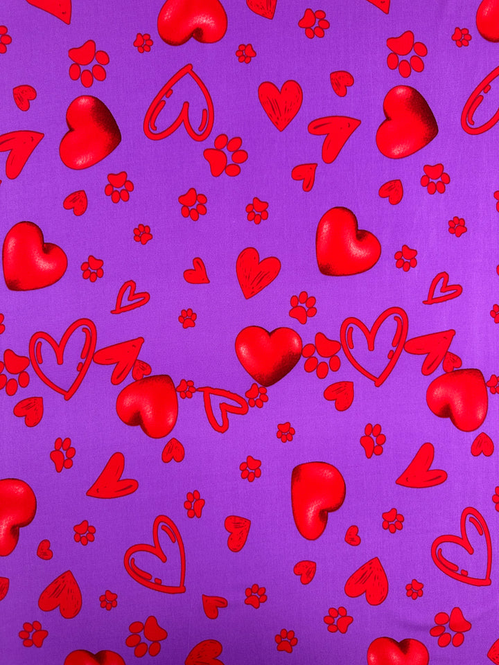 Printed Lycra - Love Riot - 150cm by Super Cheap Fabrics features a patterned purple background with scattered red hearts and small red flowers on medium weight lycra—ideal for playful, romantic spandex or polyester projects.
