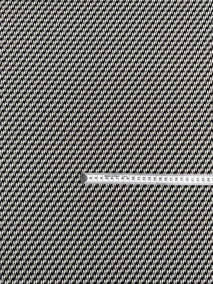 A close-up view of the Super Cheap Fabrics' Printed Lycra - Link Up (150cm) in black and white, showcasing its houndstooth pattern. A metal measuring tape is placed horizontally across the center, displaying measurements in both centimeters and inches.