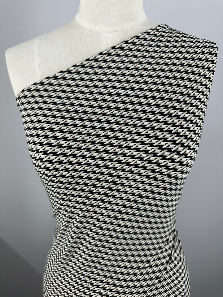 A close-up of a mannequin wearing a one-shoulder dress made from Super Cheap Fabrics' Printed Lycra - Link Up, a medium-weight, black and white houndstooth patterned fabric. The Polyester/Spandex blend wraps tightly around the form, highlighting the intricate design in a smooth, continuous flow. The background is plain and grey.