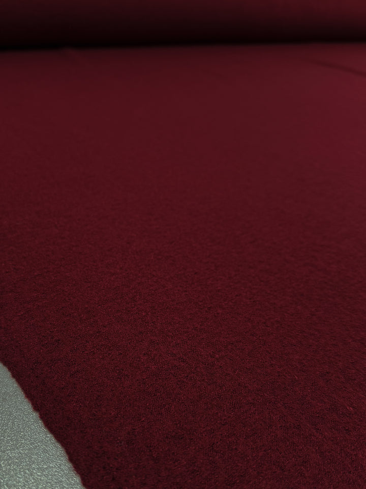 A close-up of Super Cheap Fabrics’ Boiled Wool - Rhubarb (145cm), a deep burgundy heavy weight wool with subtle texture, partially unrolled to reveal a hint of grey material at the edge.