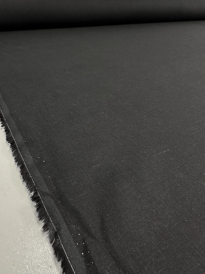 A roll of Linen Voile - Black - 140cm from Super Cheap Fabrics is unrolled across a flat surface in a well-lit room. The lightweight cotton linen appears smooth and slightly textured, extending toward the background.