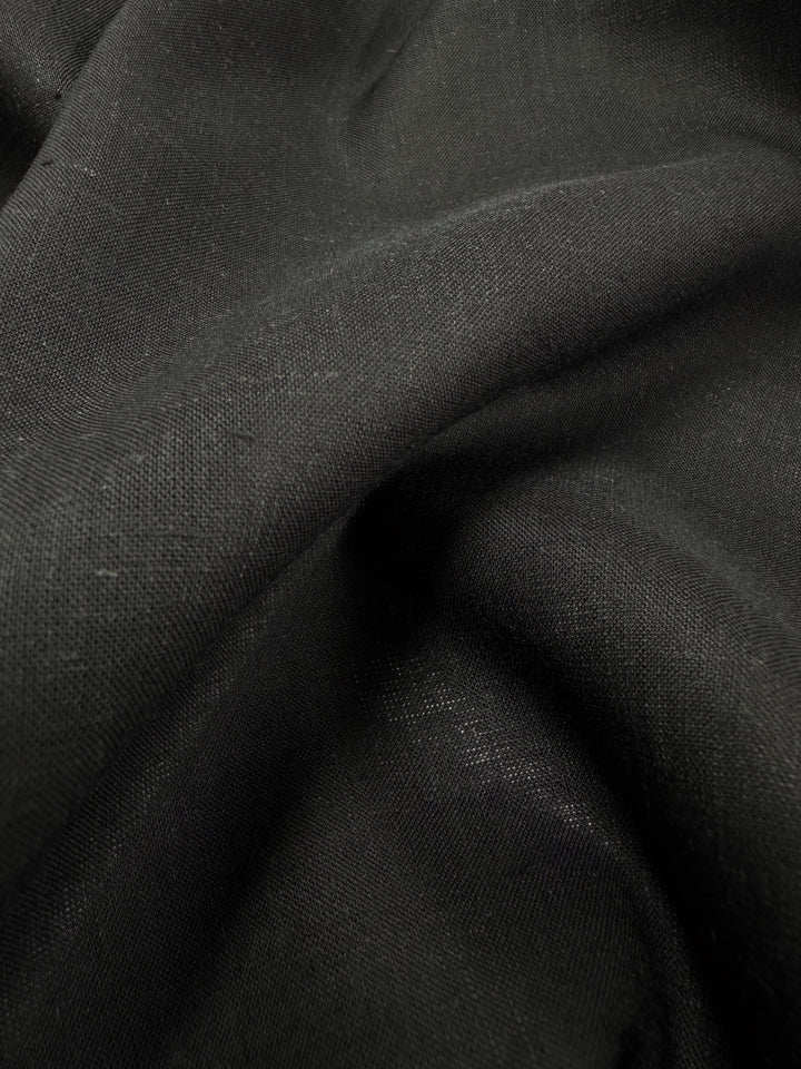 Close-up of a dark black linen voile fabric with a smooth texture and soft folds. The lighting highlights subtle variations in the material, creating depth and dimension, making it ideal for lightweight apparel. This product is known as Linen Voile - Black - 140cm by Super Cheap Fabrics.