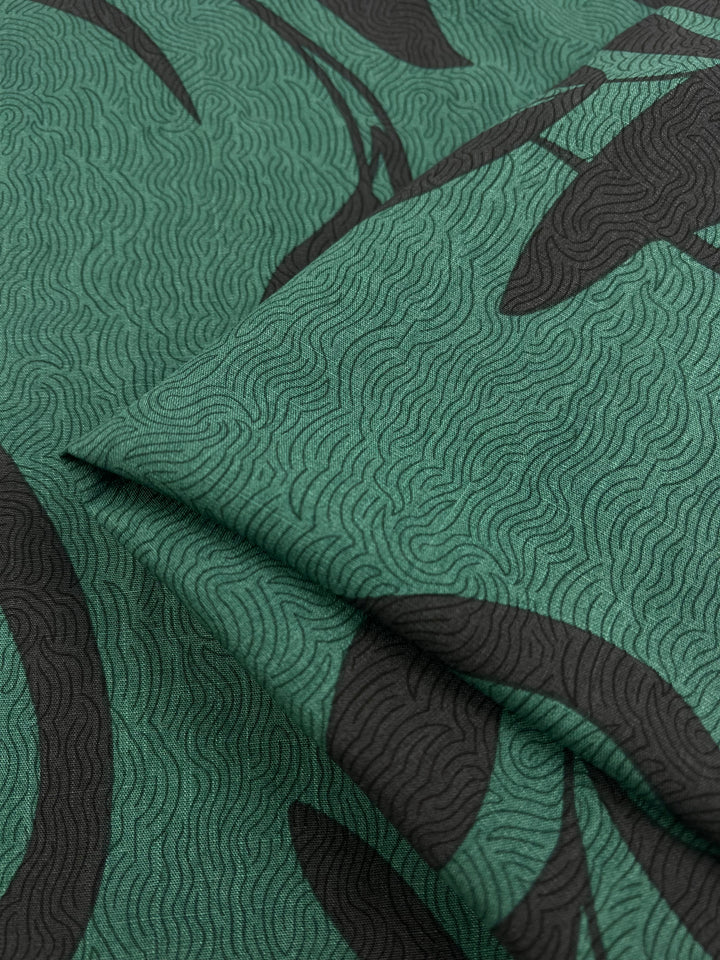 Close-up of lightweight Bamboo Rayon fabric named "Landscape" by Super Cheap Fabrics. Featuring a green and black abstract pattern, the material has fine, undulating lines on a green background with dynamic black shapes overlaying. Perfect for home decor, this 152 cm wide fabric appears folded, beautifully showcasing its texture and design.