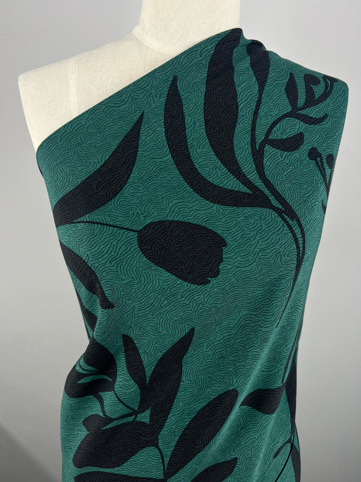 A mannequin draped with Super Cheap Fabrics' Bamboo Rayon - Landscape - 152cm, which features a dark green shade with a black floral pattern of large tulip shapes and leaves. The lightweight fabric has subtle wavy lines that create a textured look. The mannequin is displayed against a plain, light gray backdrop.