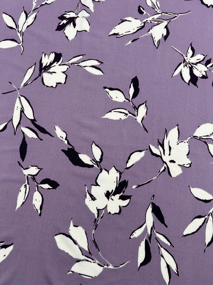 A close-up of the Printed Lycra - Dreamal - 150cm from Super Cheap Fabrics showcases its lavender background adorned with a white floral pattern. The flowers are rendered in a minimalist style, with black accents emphasizing the leaves and petals. This elegant and delicate design is ideal for medium-weight garments.