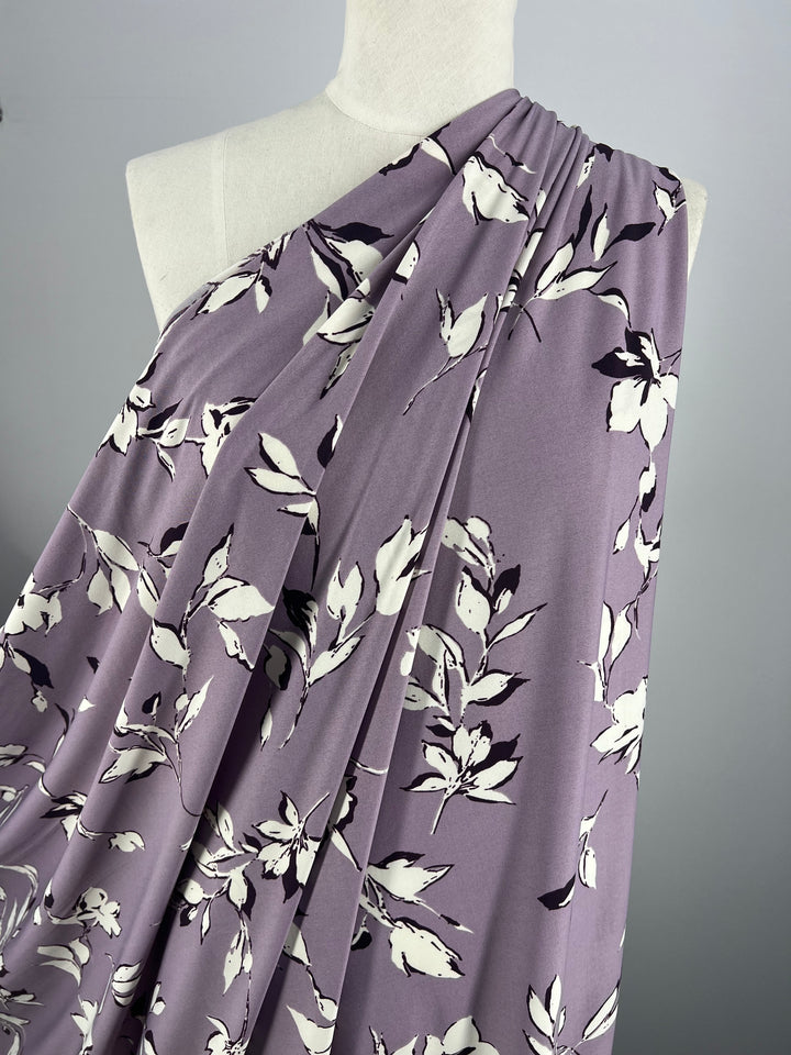 A close-up of a dress form draped with Super Cheap Fabrics' Printed Lycra - Dreamal, a 150cm wide medium-weight fabric in lavender adorned with a white floral pattern. The fabric cascades in gentle folds, highlighting its flowy texture and elegant design against a plain, light gray background.