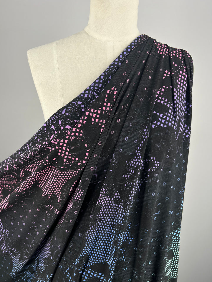 A mannequin draped in Printed Lycra - Dreamal - 150cm from Super Cheap Fabrics, featuring an intricate pattern of small dots and geometric shapes in vibrant pink, blue, and purple hues. The pattern transitions smoothly and includes subtle textures, creating a visually appealing and dynamic design.