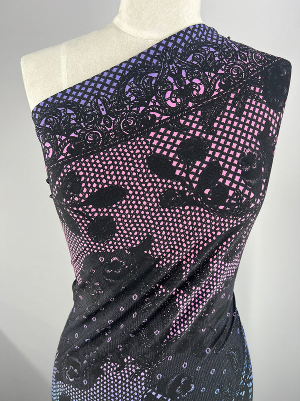 A mannequin displays a dress made from Printed Lycra - Laced - 150cm by Super Cheap Fabrics. The medium-weight Polyester/Spandex fabric showcases a black lace pattern over a gradient background that transitions from violet at the top to blue at the bottom, interspersed with small pink and white dots, in an asymmetrical one-shoulder design.