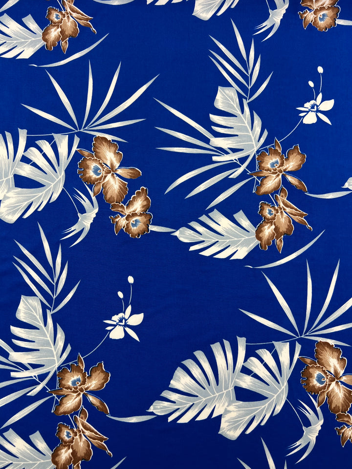 The Printed Lycra - Tropski - 150cm by Super Cheap Fabrics features a vibrant pattern of beige and brown tropical leaves and orchid-like flowers scattered across a royal blue background. Made from a polyester/spandex blend, this fabric showcases various leaf shapes and intricate floral designs, making it perfect for medium-weight projects.