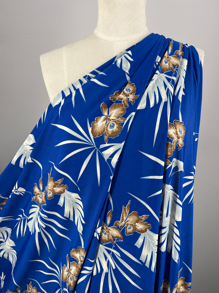 A mannequin is draped with Super Cheap Fabrics' vibrant blue medium-weight Printed Lycra - Tropski, featuring a tropical print. The pattern consists of white and beige palm leaves interspersed with brown and beige hibiscus flowers, creating a bold, summery look. The fabric measures 150cm in width.