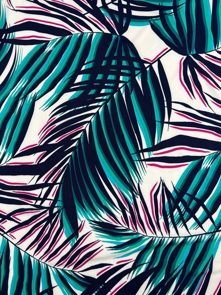 The Printed Lycra - Palma from Super Cheap Fabrics showcases a vibrant pattern of large, overlapping tropical palm leaves in dark blue, teal, and pink on a white background. The dense, intricate design creates a lively dynamic appearance, making it perfect for medium-weight polyester/spandex fabric ideal for apparel. Available in 150cm width.