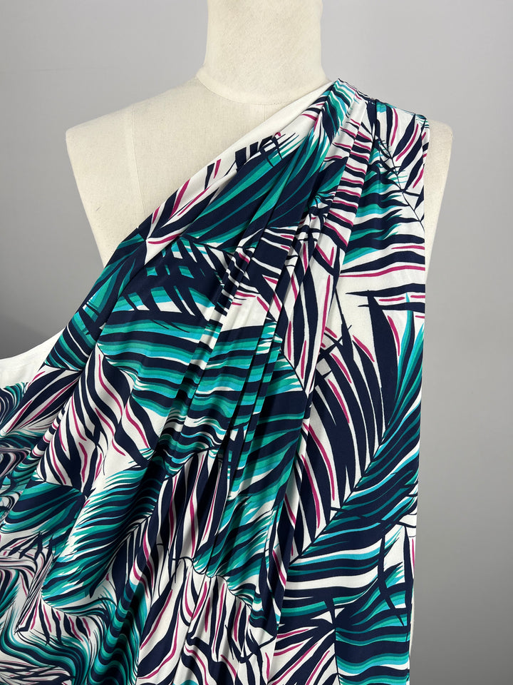 A mannequin showcases an asymmetrical dress with one sleeve, made from the medium-weight Polyester/Spandex fabric known as Printed Lycra - Palma - 150cm by Super Cheap Fabrics. The vibrant tropical leaf pattern in green, blue, and purple on a white background accentuates the garment's draped fabric, providing a flowy and elegant look.