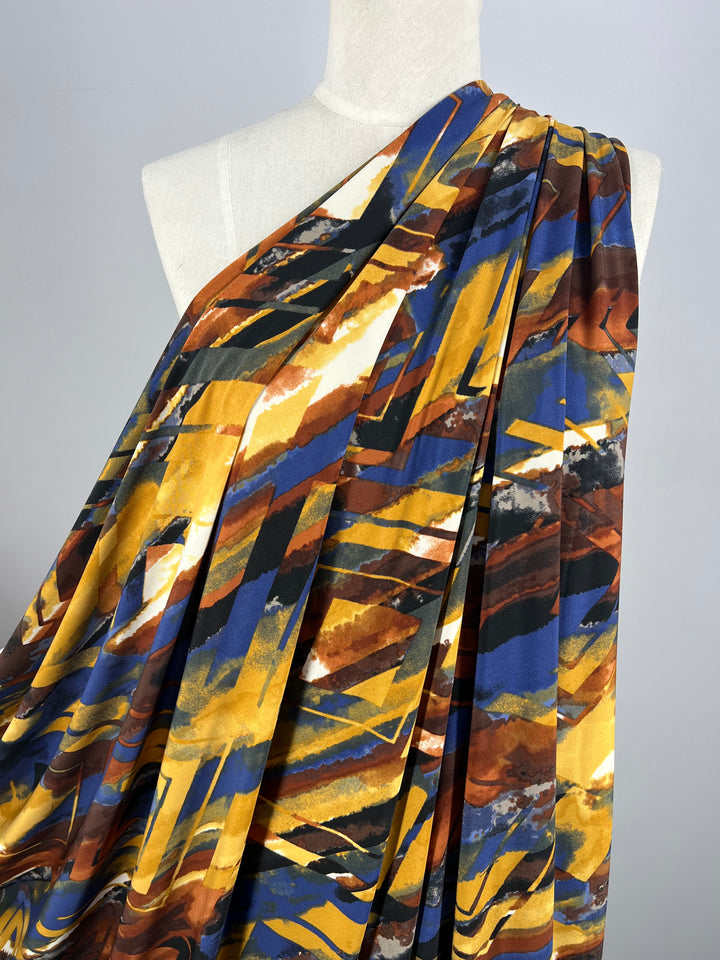 A close-up of a mannequin draped in Super Cheap Fabrics' 150cm Printed Lycra - Harlequin. The medium-weight fabric, composed of polyester and spandex, showcases a vibrant abstract pattern with bold streaks of yellow, brown, blue, and white that resemble a painted canvas. The dynamic design conveys a sense of movement and energy.