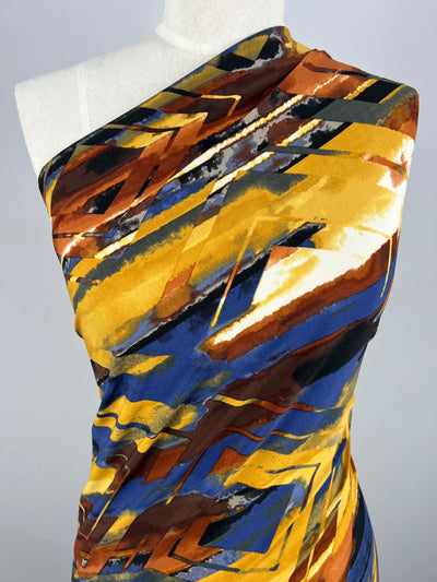 A mannequin is draped in a one-shoulder garment featuring the Printed Lycra - Harlequin by Super Cheap Fabrics. This medium-weight fabric showcases a bold, abstract design with dynamic zigzag and streaked patterns in vibrant shades of yellow, blue, brown, and white on a flexible Polyester/Spandex blend.