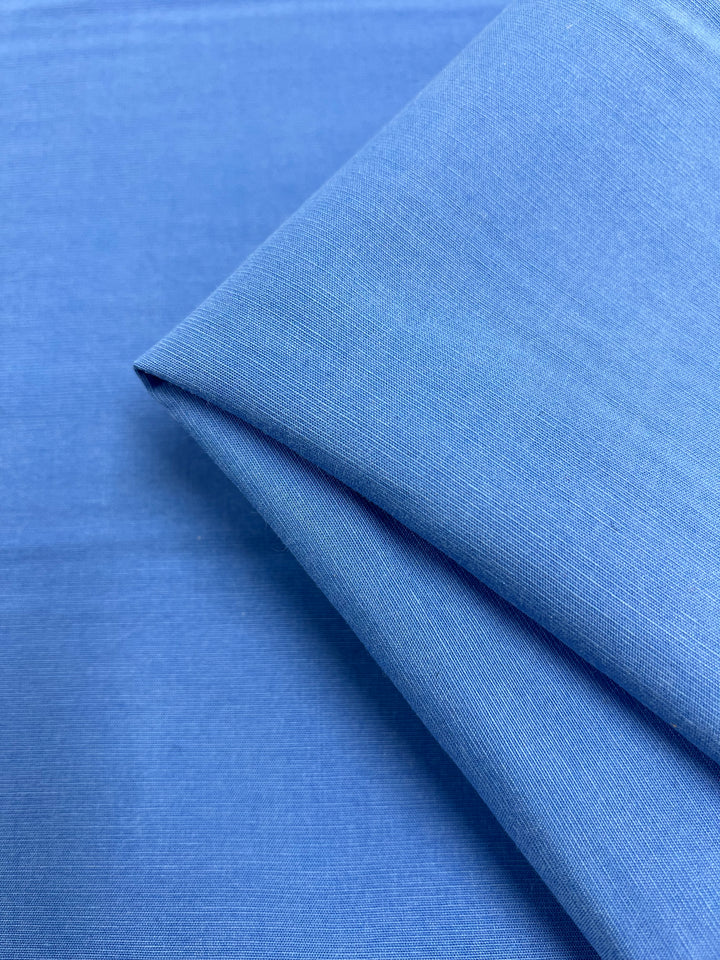 A close-up image of "Suiting - Silver Lake Blue - 145cm" fabric by Super Cheap Fabrics, neatly folded at one corner. The fabric has a smooth appearance with a subtle texture that hints at a fine weave. The color is a uniform light blue, and the neat folds suggest it might be medium weight suiting fabric made from 100% cotton.