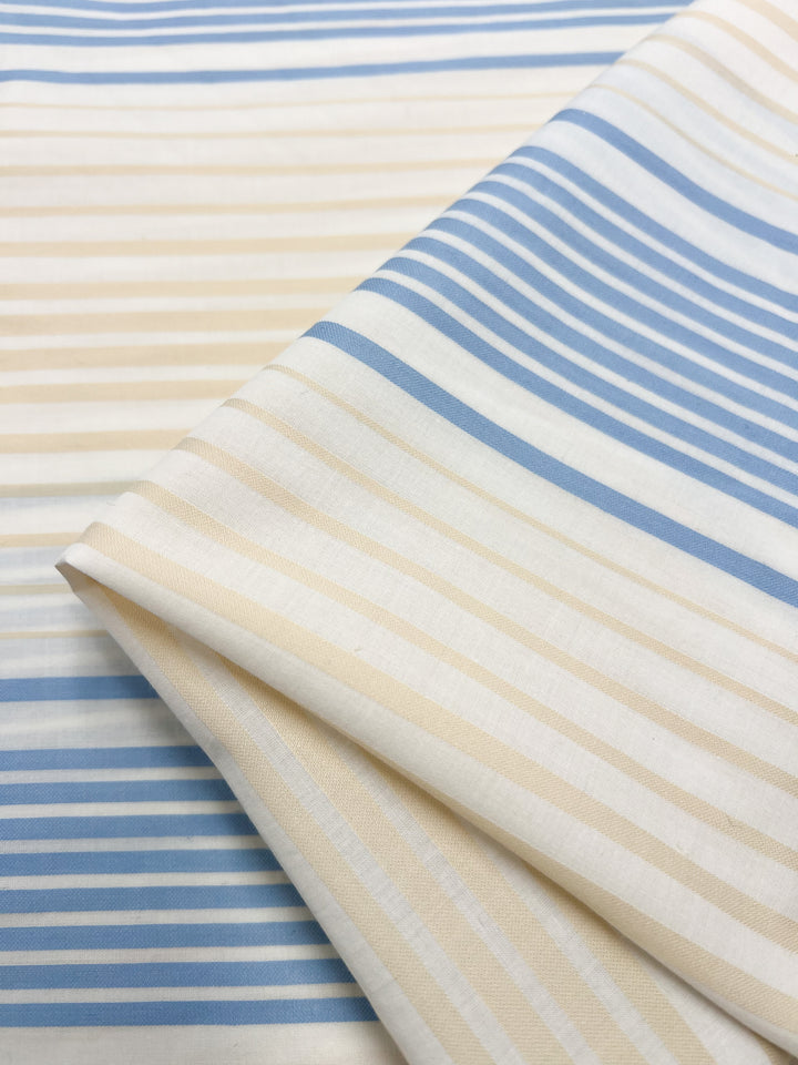 A folded piece of GOTS Cotton - Buttercream Stripe from Super Cheap Fabrics features alternating light blue and beige horizontal stripes. Made from certified GOTS organic cotton, the fabric is shown at an angle on a flat surface, highlighting its smooth texture and simple striped pattern—perfect for eco-friendly fashion.