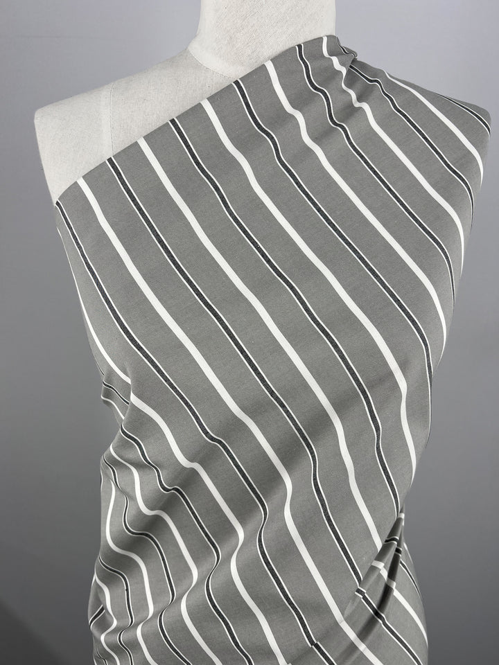 A mannequin draped in Super Cheap Fabrics' Cotton Twill - Monochrome - 153cm, featuring a single-shoulder design with diagonal white and black wavy stripes on lightweight gray fabric. The background is a simple gray gradient, emphasizing the design and texture of this cotton twill fabric.