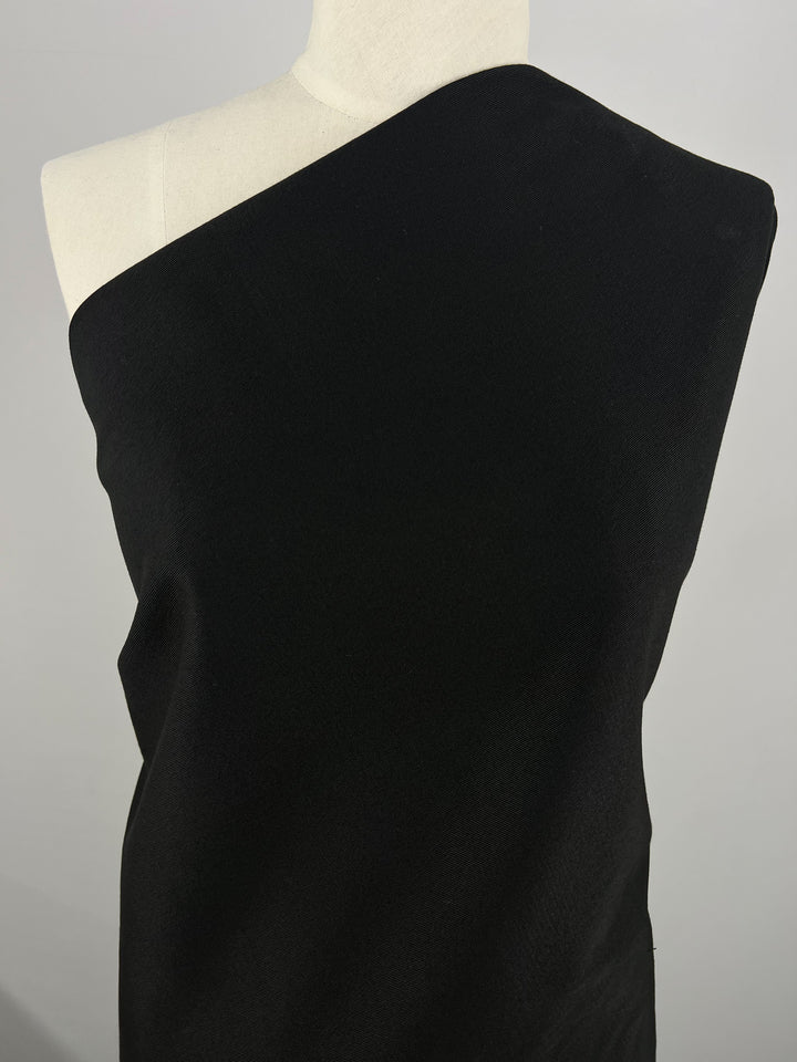 A mannequin is draped in Super Cheap Fabrics' Gabardine Suiting - Black - 140cm, a medium to heavy weight fabric. The material covers one shoulder entirely while the other shoulder remains exposed, creating an elegant, asymmetrical look. The plain gray background accentuates the showcased fabric perfectly.