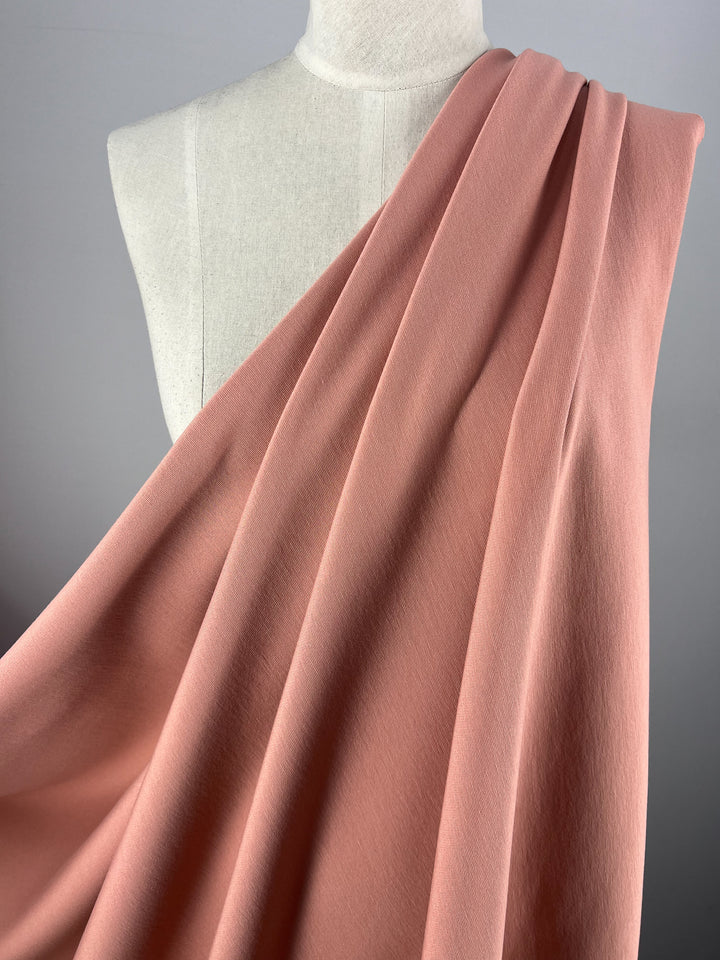 A draped Milano Ponte - Coral Cloud - 138cm fabric from Super Cheap Fabrics is displayed on a white mannequin torso, showcasing its smooth texture and elegant folds against a plain gray background.