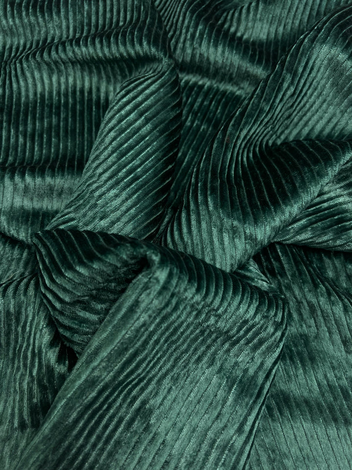 A close-up image of the Velvet Medium Cord in Forest Biome by Super Cheap Fabrics showcases its dark green, ribbed corduroy fabric with prominently visible vertical ridges and folds. This luxurious material stands out with its soft, velvety texture. The fabric width is 150cm, providing ample surface area for various sewing projects.