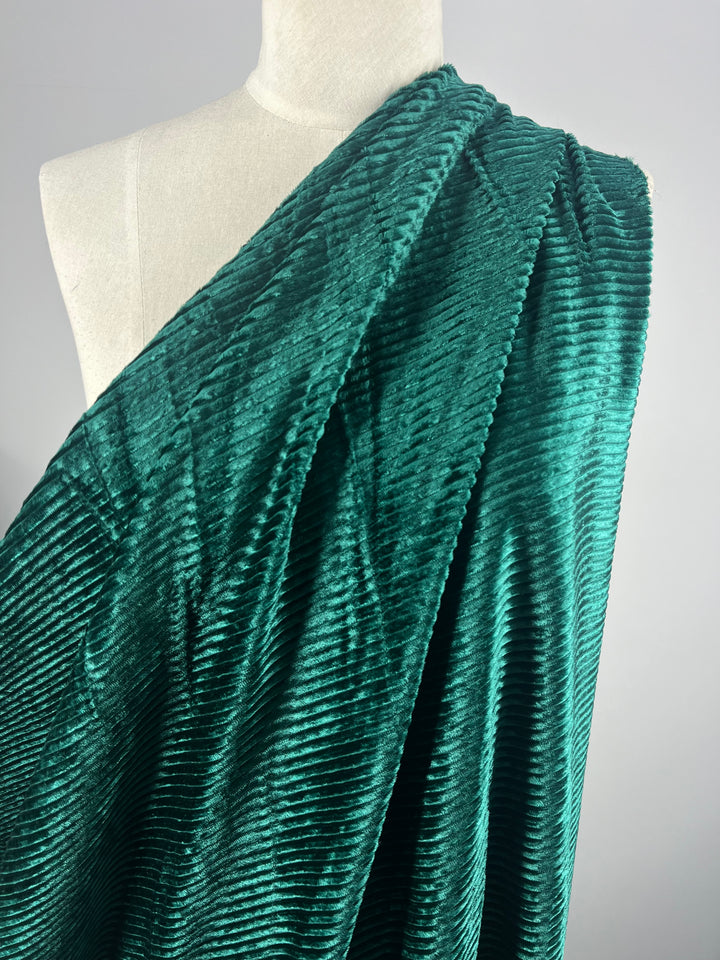 A close-up photo of a mannequin draped with Super Cheap Fabrics' Velvet Medium Cord in Forest Biome showcases the intricate, ribbed pattern and luxurious texture of this deep green, medium-weight velvet fabric. The material reflects light beautifully, highlighting its rich and opulent appearance against the plain, light-colored background that accentuates its exquisite details.