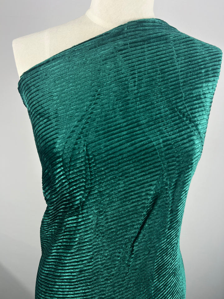 A mannequin is draped in Super Cheap Fabrics’ Velvet Medium Cord - Forest Biome - 150cm, a luxurious, emerald green heavy-weight velvet that wraps around one shoulder, creating an asymmetrical look. The fabric's ribbed, velvety appearance adds depth and dimension to the overall presentation against a plain background.