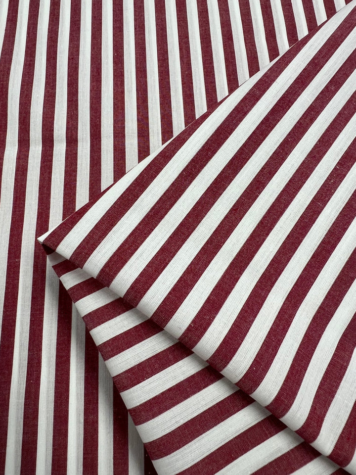 A close-up of a folded Cotton Lawn - American Beauty from Super Cheap Fabrics showcases the lightweight fabric's pattern of vertical maroon and white stripes. The neatly arranged folds highlight the consistent repetition and alignment of the stripes, making it ideal for warm weather clothing.