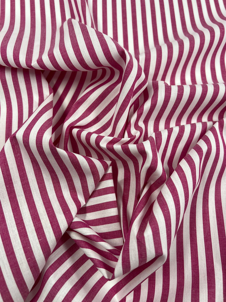 A close-up of crumpled Cotton Lawn - Lilac Rose fabric from Super Cheap Fabrics featuring vertical magenta and white stripes. The folds and creases create a dynamic pattern, giving the lightweight, 100% cotton fabric a textured appearance. The alternating stripes are of even width, contributing to a visually striking design.