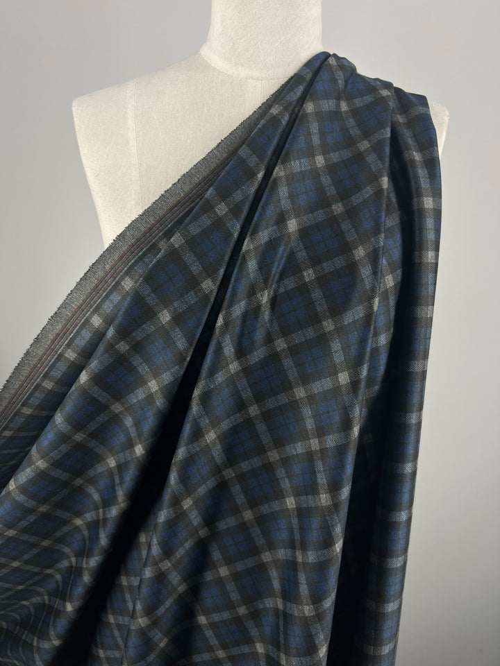 A mannequin is draped with a medium-weight piece of "Ponte - Checkers - 150cm" fabric from Super Cheap Fabrics, featuring a blue, gray, and black plaid pattern. The polyester fabric is wrapped around the mannequin's torso, showcasing its intricate pattern and texture against a plain light gray background.