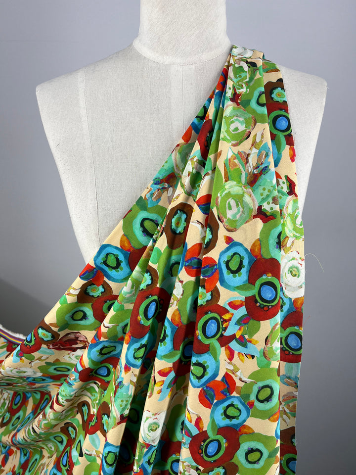 A close-up of a dress form draped in Super Cheap Fabrics' Designer Rayon - Eloise - 145cm, showcasing an abstract floral pattern with vibrant hues of green, blue, red, and brown on a beige background. The versatile fabric is artfully arranged to cover one shoulder and gathered at the waist.